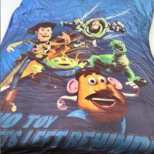 Puede incluir: Una colcha azul con personajes de la pel&iacute;cula de animaci&oacute;n "Toy Story". El dise&ntilde;o incluye a Woody, Buzz Lightyear, Rex, un extraterrestre y el Sr. Potato. El texto "NO TOY IS LEFT BEHIND" est&aacute; impreso en la parte inferior.