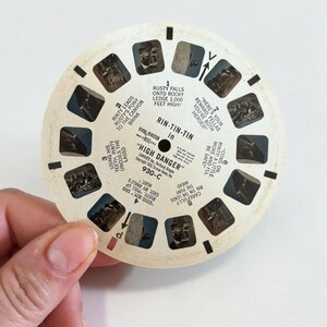 May include: A vintage View-Master reel featuring scenes from "High Danger." The circular reel has multiple small rectangular images and text, including "Rin-Tin-Tin." The reel is held by a hand.