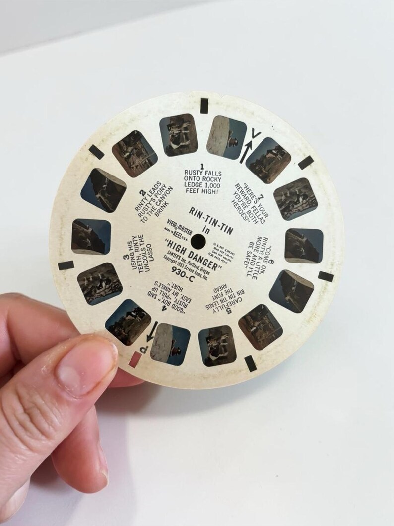 May include: A vintage View-Master reel featuring scenes from "High Danger." The circular reel has multiple small rectangular images and text, including "Rin-Tin-Tin." The reel is held by a hand.