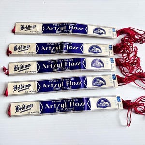 Peut inclure: Cinq échevettes de fil à broder rouge, chacune étiquetée Beldings Four Strand Artsyl Floss Boil Proof, 4 1/2 yards, numéro de teinte 528. Les étiquettes en papier sont blanches et bleues avec du texte noir et un logo circulaire représentant un chat.