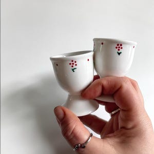 Peut inclure: Deux coquetiers en céramique blanche ornés d'un motif peint à la main composé de petits points rouges disposés en grappe florale avec une tige verte. Les coquetiers présentent une finition lisse et brillante, un bord évasé et une base stable.