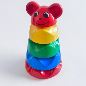 Peut inclure: Un jouet d'empilage coloré en forme de souris. Le jouet présente une tête rouge avec des oreilles et un visage souriant, empilée sur des anneaux jaunes, verts, bleus et rouges. Un jouet classique pour les jeunes enfants.