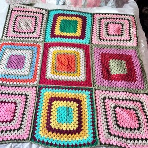 Peut inclure: Une couverture en crochet faite à la main composée d'une grille de neuf grands carrés granny. Chaque carré est formé de plusieurs rangs concentriques aux couleurs vives, notamment rose, jaune, vert, orange, rouge, bleu et violet, assemblés par un fil vert foncé.