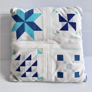 May include: Square white cushion with a cross-stitch design. The cushion features four sections, each with a different geometric pattern in shades of blue and teal. The stitching is detailed, and the cushion has a soft, textured appearance.