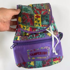 May include: A purple backpack featuring a multi-colored patchwork pattern of flowers, frogs, and castles on the flap and base. The front pocket has a zipper closure and the text "CAMPERINO FOR HER" printed in red, surrounded by decorative hearts.