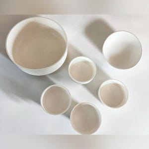 Peut inclure: Un ensemble de six bols en céramique blanche de tailles variées disposés en groupe. La collection comprend un grand bol, un bol moyen et quatre bols plus petits, tous dotés d'une finition lisse et mate et de designs circulaires ouverts.