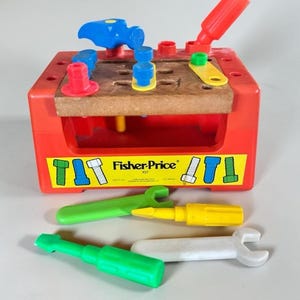 May include: A red plastic toy workbench with a brown top featuring holes for plastic bolts and pegs. The front displays a yellow Fisher-Price 927 label. Included are a blue hammer, red screwdriver, green wrench, yellow screwdriver, and white wrench.