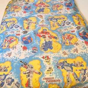 May include: A light blue bed sheet featuring a repeating pattern of Transformers characters and vehicles. The design includes Optimus Prime and other robots in yellow clouds, with the text Transformers printed in red and black near the bottom.