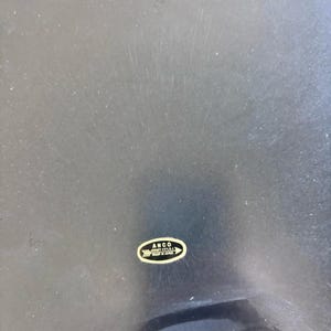 May include: A dark, possibly black, surface with visible scratches. A small, oval-shaped sticker in gold and white is affixed to the surface, bearing the text "ANCO JERSEY CITY N.J. MADE IN JAPAN".