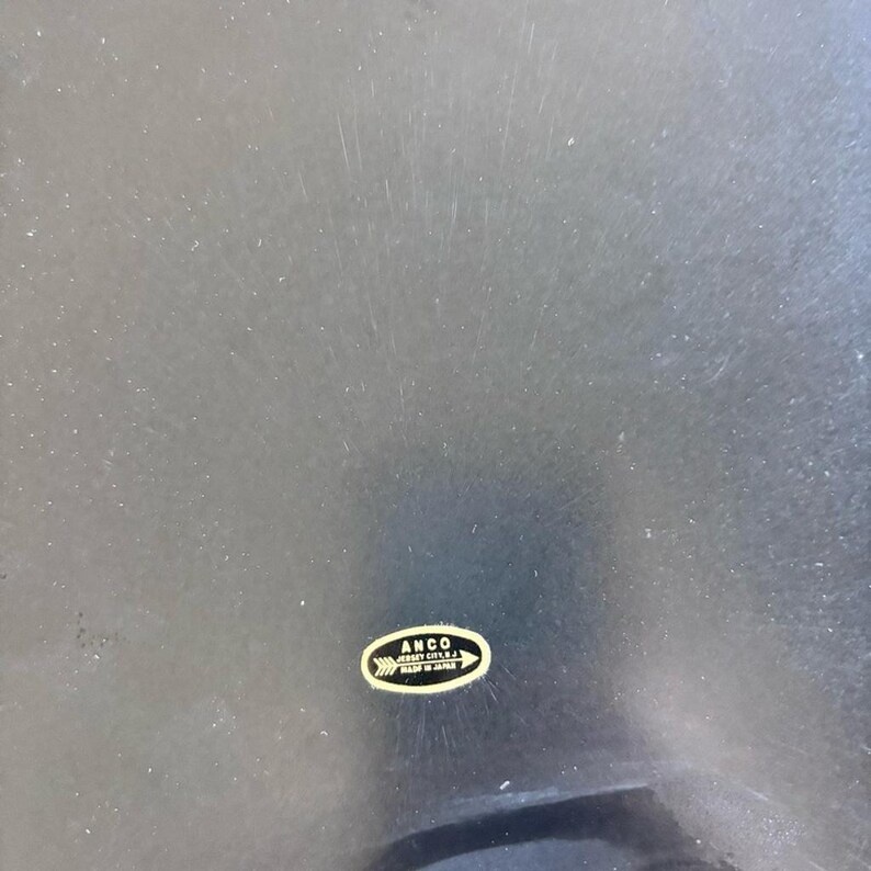 May include: A dark, possibly black, surface with visible scratches. A small, oval-shaped sticker in gold and white is affixed to the surface, bearing the text "ANCO JERSEY CITY N.J. MADE IN JAPAN".