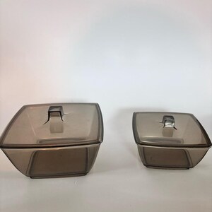 Peut inclure: Deux récipients carrés avec couvercles en verre translucide de couleur fumée. Chaque récipient présente une base effilée et un couvercle assorti avec une petite poignée carrée centrée. Les récipients sont placés côte à côte, l'un plus grand.