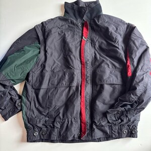 May include: A black jacket featuring a high collar, long sleeves with buttoned cuffs, and a front zipper closure concealed by a placket. The garment includes dark green panels on the sleeves and a vibrant red interior lining visible along the front opening.