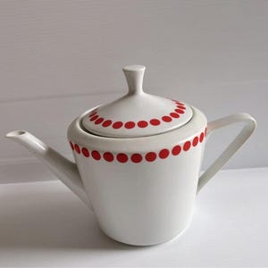Peut inclure: Une théière en céramique blanche avec un corps conique, un couvercle assorti muni d'un bouton de préhension et un bec verseur incurvé. La théière est ornée d'une rangée horizontale de petits pois rouge vif sur le couvercle et le bord supérieur.