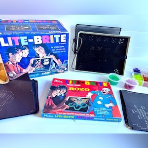 May include: A vintage Lite-Brite toy set with boxes and accessories. The main box is blue with the Lite-Brite logo and images of children using the toy. Additional boxes and a light-up screen are also visible, along with colorful pegs.