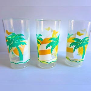 Peut inclure: Trois verres transparents ornés d'un motif graphique tropical. Chaque verre présente un design coloré composé de palmiers verts, de soleils jaunes et de voiles de bateau blanches à rayures orange sur un corps en verre transparent.
