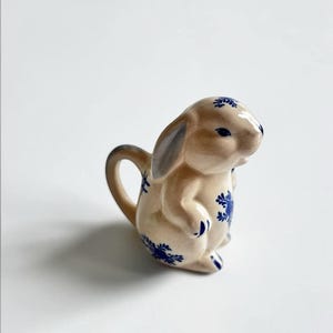 Peut inclure: Une crémière en céramique en forme de lapin, avec un corps beige et des motifs floraux bleus. Le lapin a une anse et un bec verseur, et ses oreilles sont d'une teinte plus foncée. Objet décoratif.