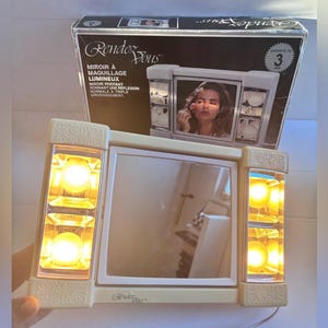 Peut inclure: Un miroir de maquillage lumineux Rendez-Vous beige avec une vitre centrale rectangulaire et deux panneaux latéraux, chacun contenant deux ampoules allumées. Le produit est présenté devant sa boîte d'emballage en carton avec du texte.