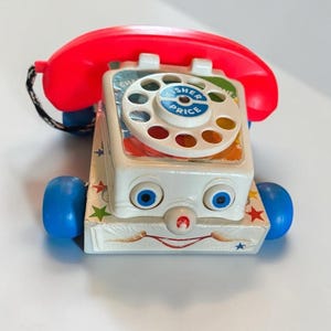 May include: A vintage Fisher-Price pull-toy telephone with a white body, a bright red receiver, and blue wheels. The front features a painted face with blue eyes and a red nose, while the rotary dial has the Fisher-Price logo in the center.