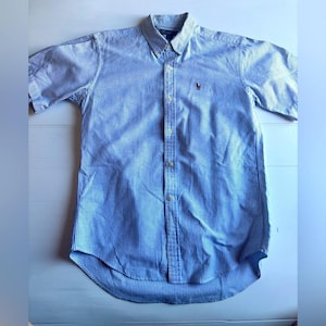 May include: A light blue and white striped button-down shirt with short sleeves. The shirt has a button-down collar and a small embroidered logo on the chest. The shirt is made of a lightweight fabric.