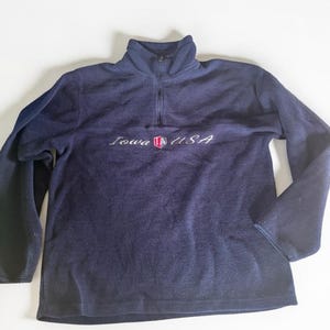Peut inclure: Un pull en polaire bleu marine à fermeture éclair quart, doté d'un col montant et de manches longues. La poitrine présente le texte brodé Iowa USA en écriture blanche, centré avec un petit écusson rouge, blanc et bleu portant la lettre A.