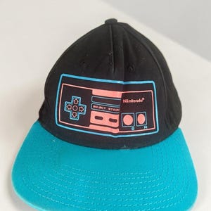 Könnte beinhalten: Eine schwarze Baseballkappe mit einem blaugrünen Schirm. Auf der Vorderseite befindet sich eine Grafik eines Retro-Videospiel-Controllers in Hellblau und Pink, mit dem in Pink gedruckten Wort Nintendo. Das Design zeigt ein Steuerkreuz.
