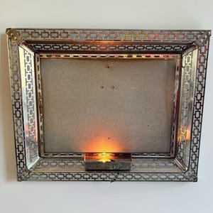 Peut inclure: Un cadre photo rectangulaire en métal avec une finition argentée et un motif géométrique en treillis. Le cadre comporte un luminaire central sur le bord inférieur qui émet une lueur orange chaude, éclairant le fond en carton.