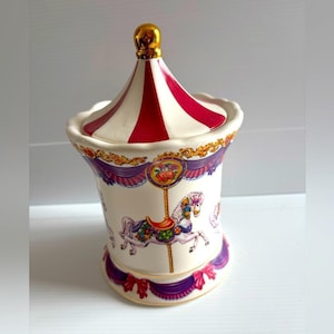 Op de afbeelding: Een keramische pot met carrouselthema en een deksel. De pot heeft een rood-wit gestreepte bovenkant, een gouden knop en een basis versierd met carrouselpaarden en bloemaccenten. De pot is ongeveer 15 cm hoog.