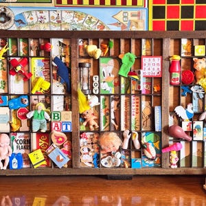 May include: A wooden printer's tray divided into numerous small rectangular compartments, each filled with a variety of miniature toys, figurines, colourful blocks, and small printed cards. The word GAMES is spelled out in red blocks at the top right.
