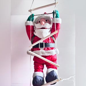 May include: A plush Santa Claus figure climbing a rope ladder with wooden rungs. The figure wears a shiny red suit with white faux fur trim, green gloves, a black belt with a silver buckle, and dark boots. The Santa has a white beard and hat.