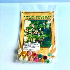 May include: A Christmas ornament set in a clear plastic bag featuring a yellow cardboard insert with the text Christmas Ornament Set and Ornements de Noël. The kit includes small paint pots labeled with numbers and a printed image of ornaments.