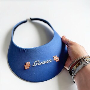 May include: A blue sun visor featuring the word Guess embroidered in white thread across the center, flanked by two small square geometric patterns in light orange and white thread on either side of the brand name.
