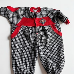 May include: A long-sleeved one-piece jumpsuit featuring a black and white chequered pattern with red fabric accents on the shoulders, chest, and cuffs. The garment includes a front button closure and a diamond-shaped patch on the chest with text.