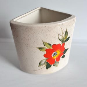 Peut inclure: Un vase en céramique beige avec une finition mouchetée et un sommet triangulaire. Le vase est orné d'une fleur rouge vif avec un centre jaune et des feuilles vertes. Le vase est probablement à des fins décoratives.