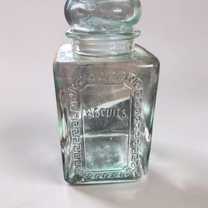 Peut inclure: Un bocal en verre transparent de couleur vert clair avec un couvercle en verre assorti. Le récipient de forme carrée présente un motif de bordure grecque en relief entourant le mot BISCUITS, moulé au centre de la face avant du verre.