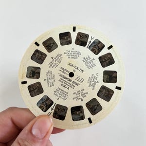 May include: A vintage View-Master reel featuring scenes from "Vanishing Guns." The circular reel has multiple small rectangular windows with images and text. The text includes "Rin-Tin-Tin" and scene descriptions. The reel is held by a person.