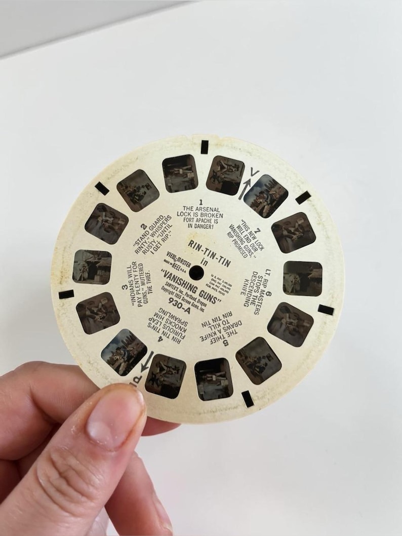 May include: A vintage View-Master reel featuring scenes from "Vanishing Guns." The circular reel has multiple small rectangular windows with images and text. The text includes "Rin-Tin-Tin" and scene descriptions. The reel is held by a person.