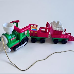May include: A Fisher-Price pull toy train consisting of a green engine with a white smokestack, a red open-top cargo car containing a small block, and a red caboose with a white roof. A long tan string is attached to the front of the engine.