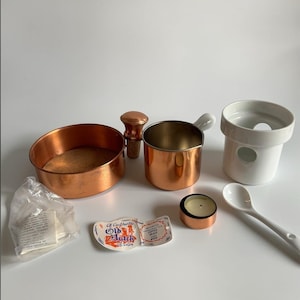 Pode incluir: Uma coleção de utensílios de cozinha de cobre, incluindo uma panela pequena com uma alça branca, uma panela redonda de cobre, um recipiente de cerâmica branco, uma vela pequena e uma colher branca. Também inclui uma rolha de cortiça e uma etiqueta que diz "Old Dutch Design".
