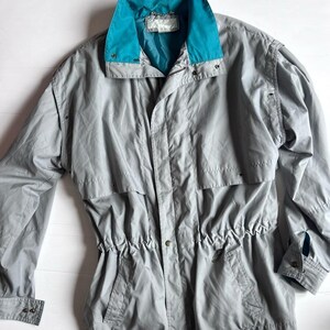 May include: A light gray windbreaker jacket featuring a vibrant teal collar, a front zipper closure with a snap-button placket, long sleeves with adjustable cuffs, and a drawstring waist. The jacket includes two front pockets and a horizontal seam.