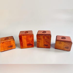 Puede incluir: Cuatro recipientes de almacenamiento cuadrados de madera con tapas, cada uno con una pequeña abertura ovalada en la parte superior. Los recipientes tienen un acabado marrón cálido y etiquetas con texto como Coffee, Sugar y Tea, alineados.