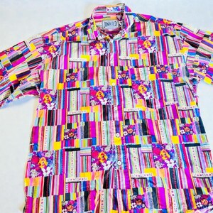 Peut inclure: Chemise à manches courtes avec un motif patchwork aux couleurs vives. Le motif comprend des rayures verticales et des motifs floraux dans un mélange de rose, jaune et bleu. La chemise a un col et la marque "JUNIOR D" est visible.