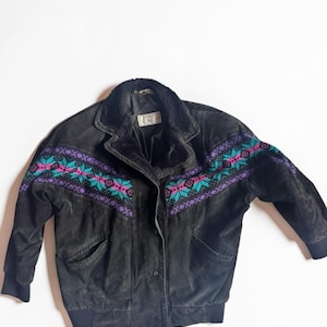 Peut inclure: Une veste en daim noir avec un col en fausse fourrure noire et une bande horizontale sur la poitrine et les manches ornée d'un motif géométrique dans des tons turquoise, magenta et violet. La veste possède une fermeture à boutons et des poignets côtelés noirs.