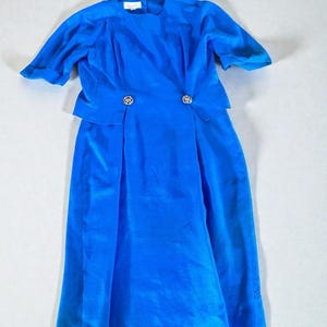 Peut inclure: Une robe bleu roi vif avec des manches courtes, une encolure ronde et un corsage structuré. La taille est ornée de deux boutons circulaires argentés. La jupe tombe à une longueur midi avec une silhouette droite aux lignes épurées.