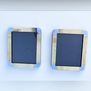 Peut inclure: Deux cadres photo rectangulaires dotés d'une bordure métallique dorée avec une texture finement nervurée verticalement. Les coins de chaque cadre sont accentués par des plaques métalliques argentées lisses et polies, entourant un centre sombre.