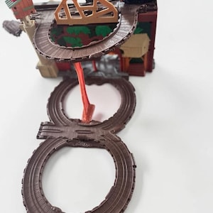 May include: A plastic toy train set featuring a brown figure-eight track layout, a small blue locomotive, a tan bridge structure, and a green and red building. The elevated track is supported by a red pillar, creating a multi-level railway system.