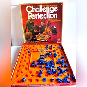 Peut inclure: Jeu de société Challenge Perfection de Lakeside avec une boîte rouge illustrée d'enfants jouant. Le jeu comprend un plateau en plastique orange avec des découpes géométriques, des pièces bleues et une figurine en forme de trophée.