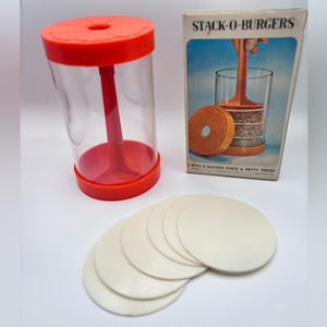 Peut inclure: Un kit vintage "Stack-O-Burgers" comprenant une presse à burger en plastique rouge et transparent, six disques séparateurs blancs et la boîte d'origine. La boîte présente une illustration de la presse à burger en cours d'utilisation. Le kit est conçu pour faire des burgers empilés.