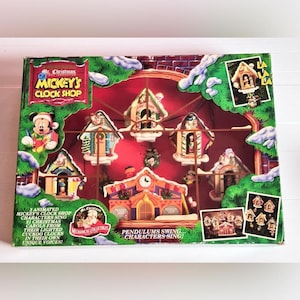 May include: A Mr. Christmas Mickey's Clock Shop mechanical collectible set in its original box. The packaging features a red background with five miniature cuckoo clock houses, each containing a Disney character, and a central clock building.