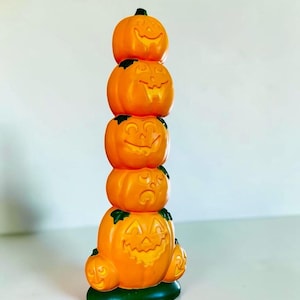 Peut inclure: Une pile verticale de cinq citrouilles orange en forme de jack-o'-lantern avec des visages sculptés, dotées de tiges et d'accents verts, reposant sur une base circulaire verte solide. Les citrouilles varient en taille, de la plus grande à la base à la plus petite au sommet.