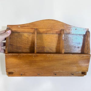 Peut inclure: Un organisateur mural en bois avec une finition en grain de bois brun clair naturel. L'unité comprend deux compartiments supérieurs pour le courrier ou les documents et une étagère horizontale inférieure avec quatre petits crochets en métal.